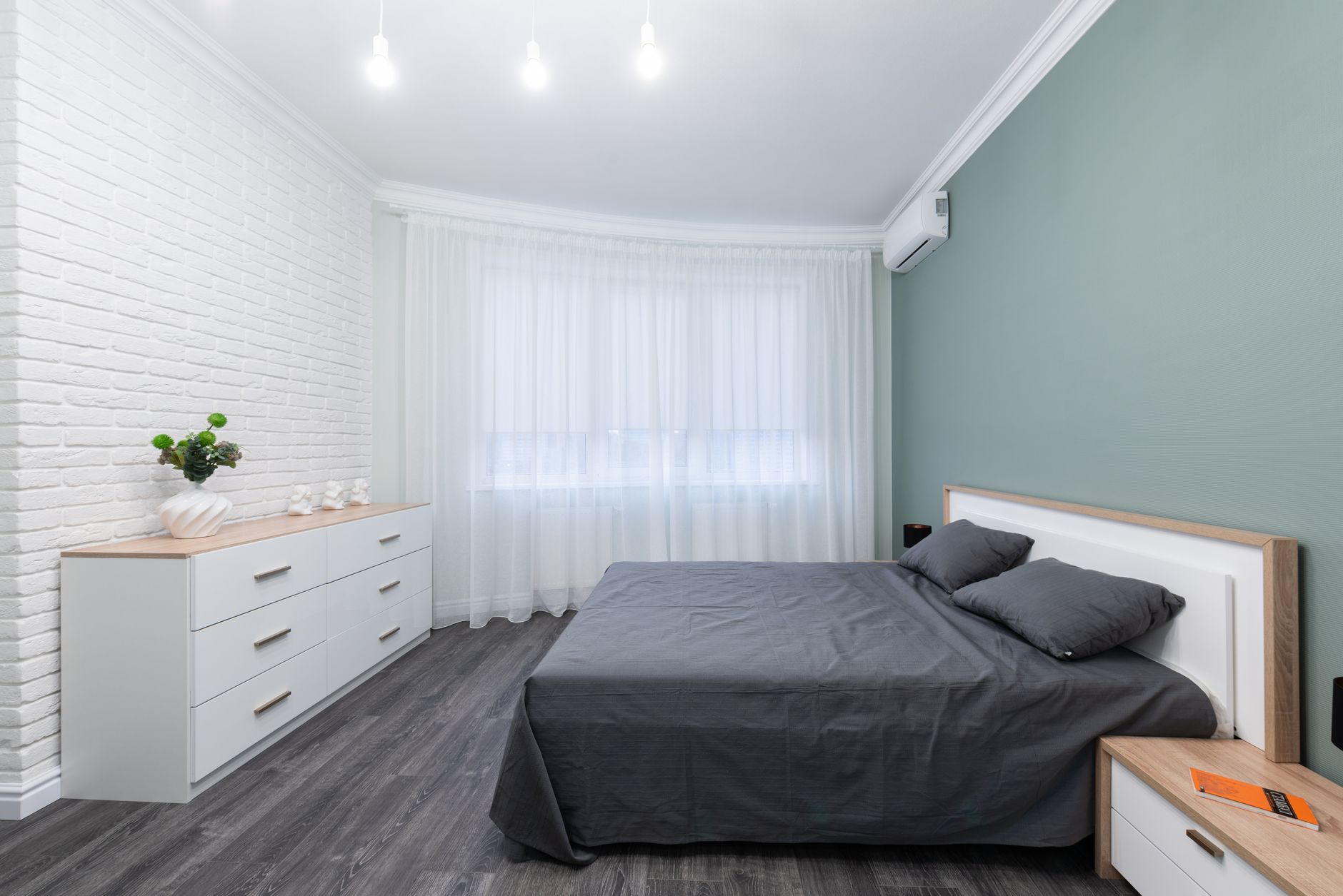 Chambre claire avec lit à literie grise, commode blanche, mur de briques blanches, voilage à la fenêtre et mur vert grisé.