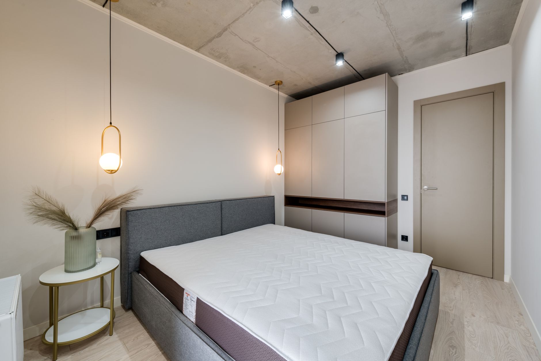 Chambre moderne avec un lit gris au matelas blanc, une armoire haute beige jusqu’au plafond, une table d’appoint ronde et deux suspensions.