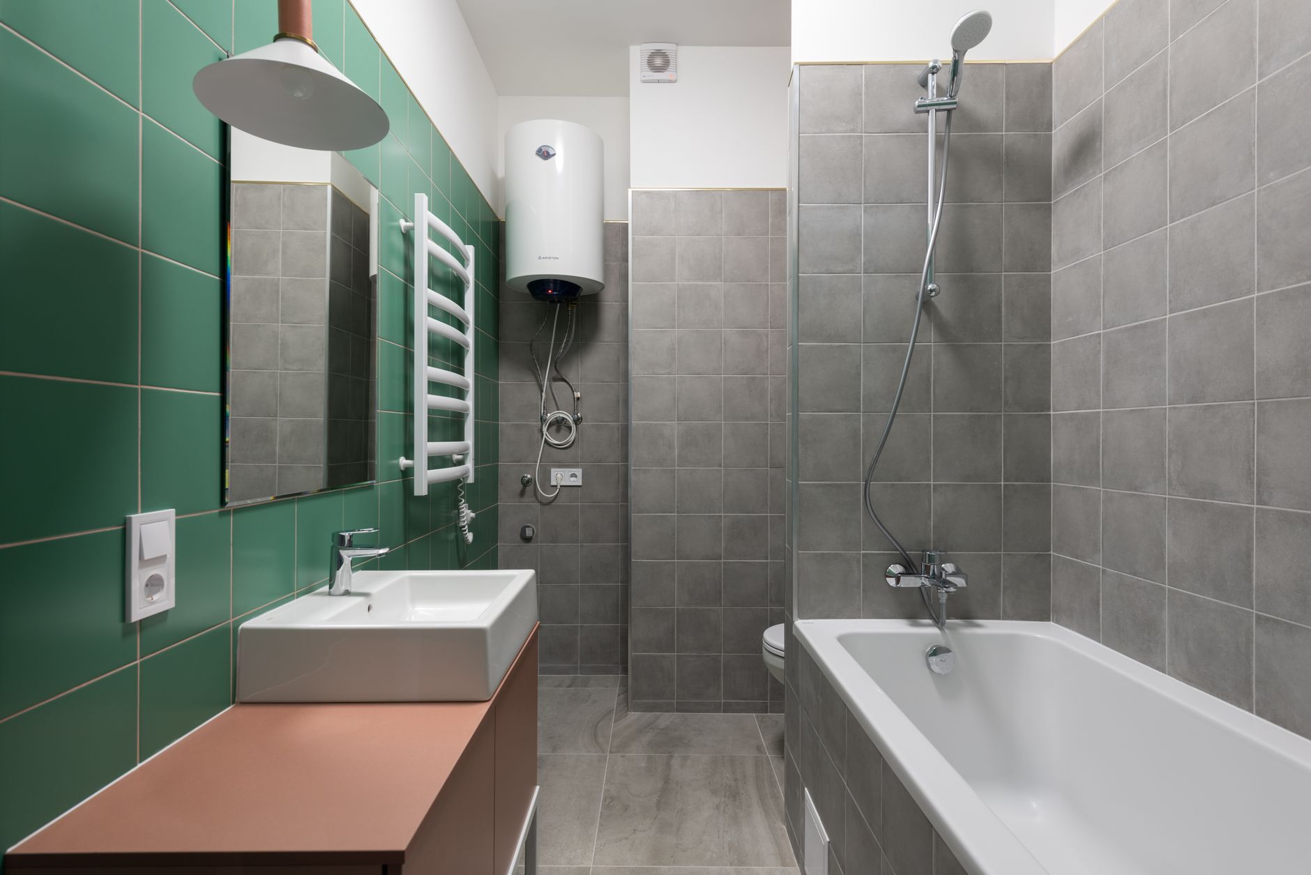 Salle de bains avec carrelage mural vert et gris, vasque blanche sur meuble terracotta, sèche-serviettes, chauffe-eau et baignoire.