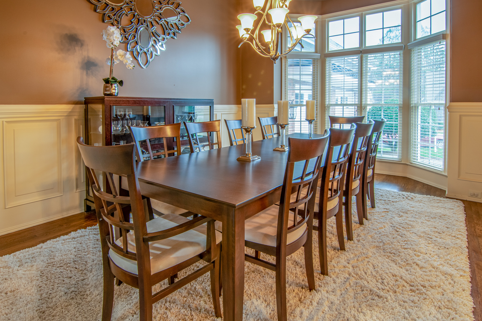 Salle à manger avec table rectangulaire en bois foncé, chaises assorties, bougeoirs, tapis beige, vitrine et grandes fenêtres.