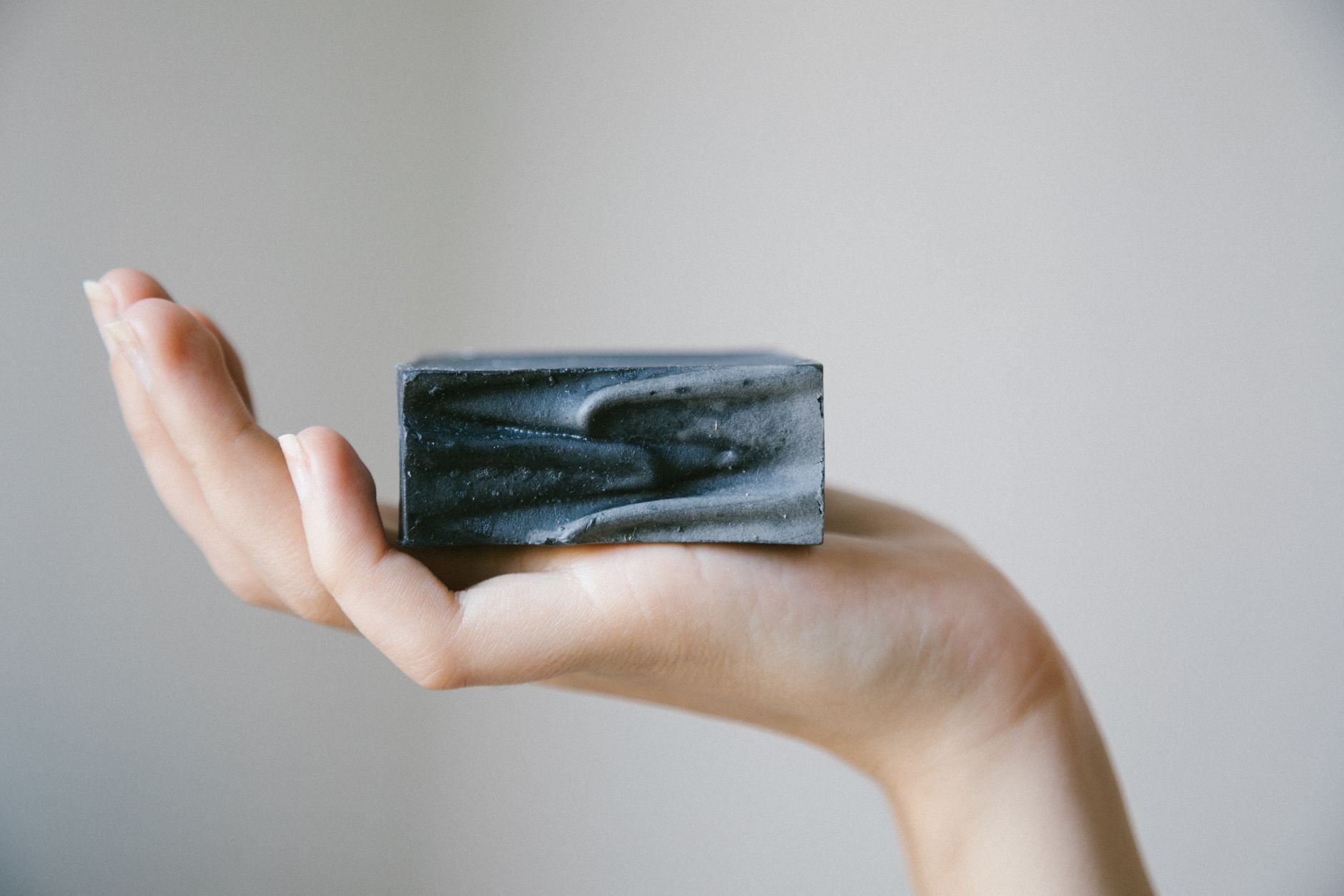 Une main tient un pain de savon rectangulaire gris foncé à la surface ondulée, devant un fond beige uni.